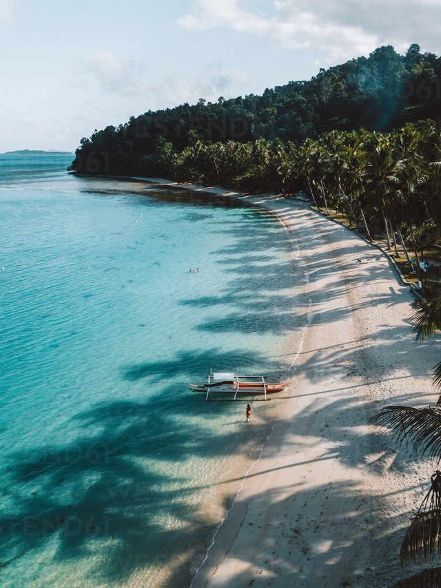 Una vista aerea di una spiaggia tropicale con una canoa a bilanciere in acqua turchese e lunghe ombre di palme sulla sabbia bianca.