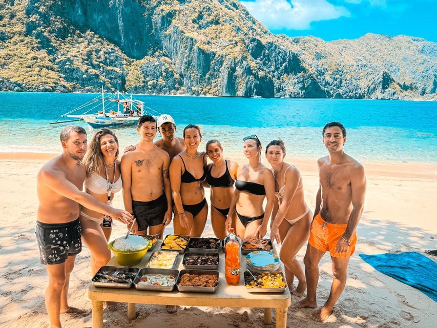 Un gruppo WeRoad in viaggio si raduna attorno a un tavolo imbandito su una spiaggia sabbiosa, con scogliere rocciose e una barca sullo sfondo.