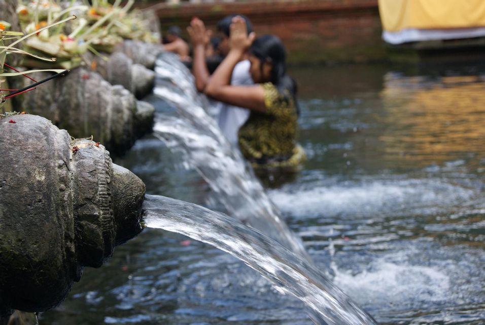 L'acqua scorre da fontane di pietra scolpite in una vasca, dove sullo sfondo sfocato le persone compiono un rituale di purificazione.