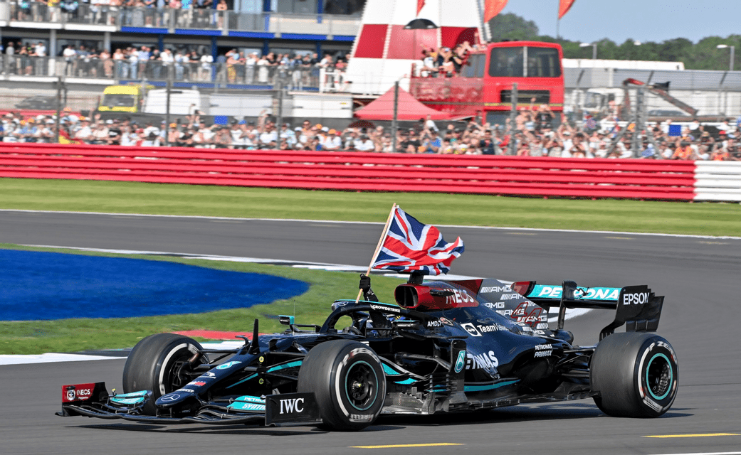 A Formula 1 driver in a black race car holds up a British flag while driving on a racetrack in front of spectators.