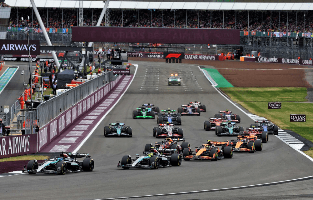A fleet of Formula 1 cars racing in a pack on a circuit with a crowded grandstand in the background.