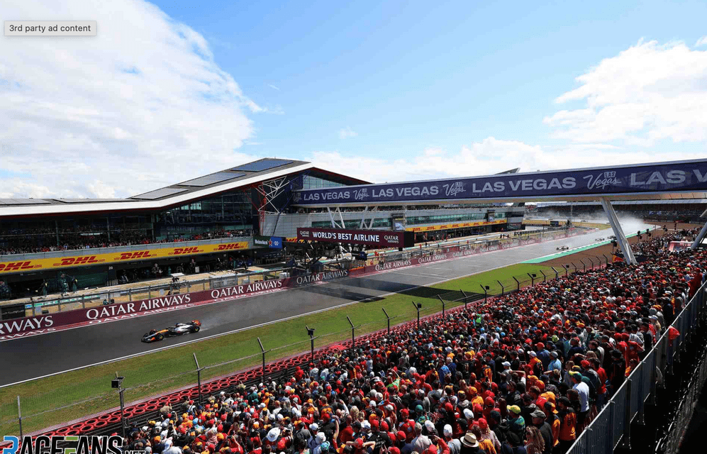 A wide shot of a race track with a single car speeding past a grandstand packed with spectators under a blue sky.