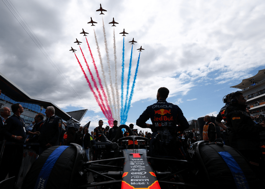 From behind a Formula 1 car, a driver and crew watch jets perform a flyover, leaving trails of red, white, and blue smoke.