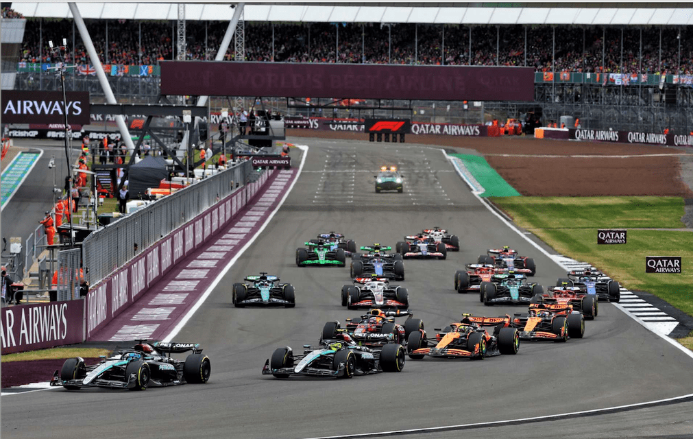 A pack of Formula 1 cars race down a track with grandstands full of spectators in the background.