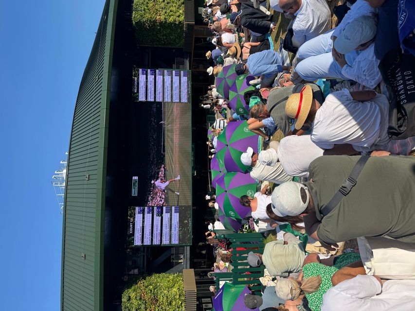A crowd of spectators sitting on a grassy hill watch a live tennis match on a large outdoor screen, some using umbrellas for shade.