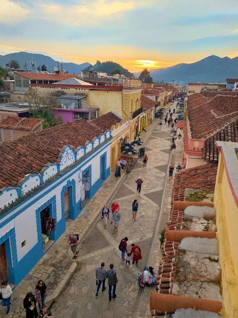 An elevated view of people walking down a narrow street in a colorful town at sunset, with mountains visible in the distance.