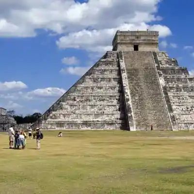 A WeRoad group trip on a grassy field looking at a large stone step-pyramid under a partly cloudy sky.