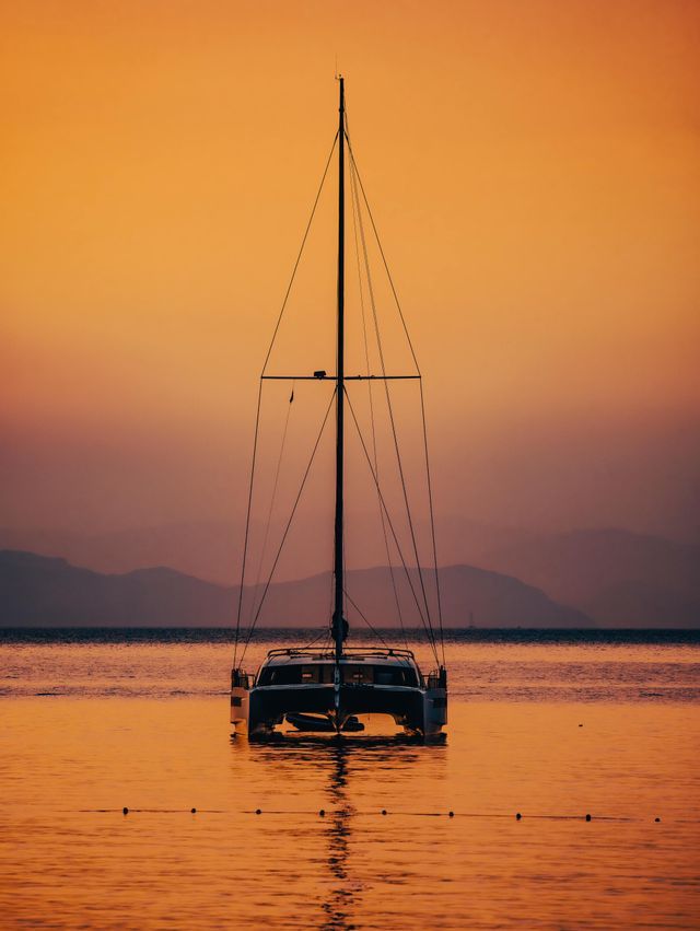 Un voilier catamaran ancré sur une eau calme, sous un ciel de coucher de soleil orange et des montagnes lointaines.