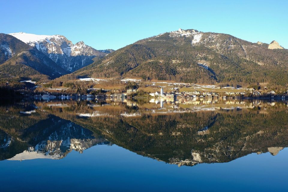 Un villaggio in riva al lago e montagne innevate si riflettono perfettamente nelle acque calme e blu del lago in una giornata di sole.