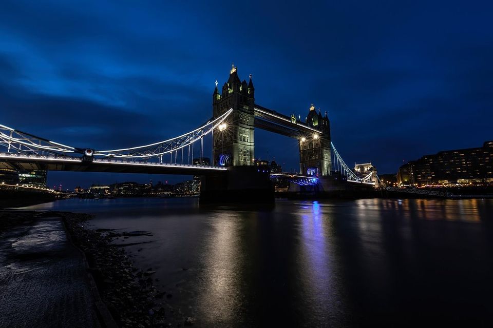 Eine große, beleuchtete Brücke mit zwei Türmen steht nachts über einem Fluss, ihre Lichter spiegeln sich auf der Wasseroberfläche wider.