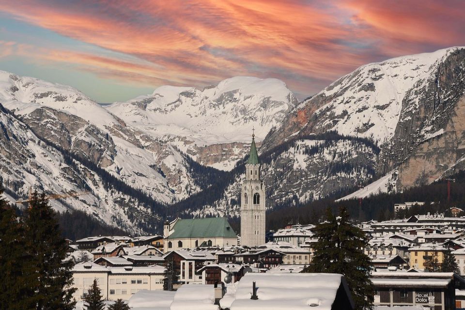 Un villaggio innevato con un'imponente torre di chiesa alla base di grandi montagne coperte di neve sotto un cielo drammatico rosa e arancione.