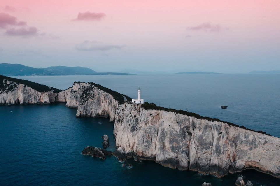 Un faro bianco si erge su una lunga penisola rocciosa circondata dal mare sotto un cielo rosa e grigio al tramonto.