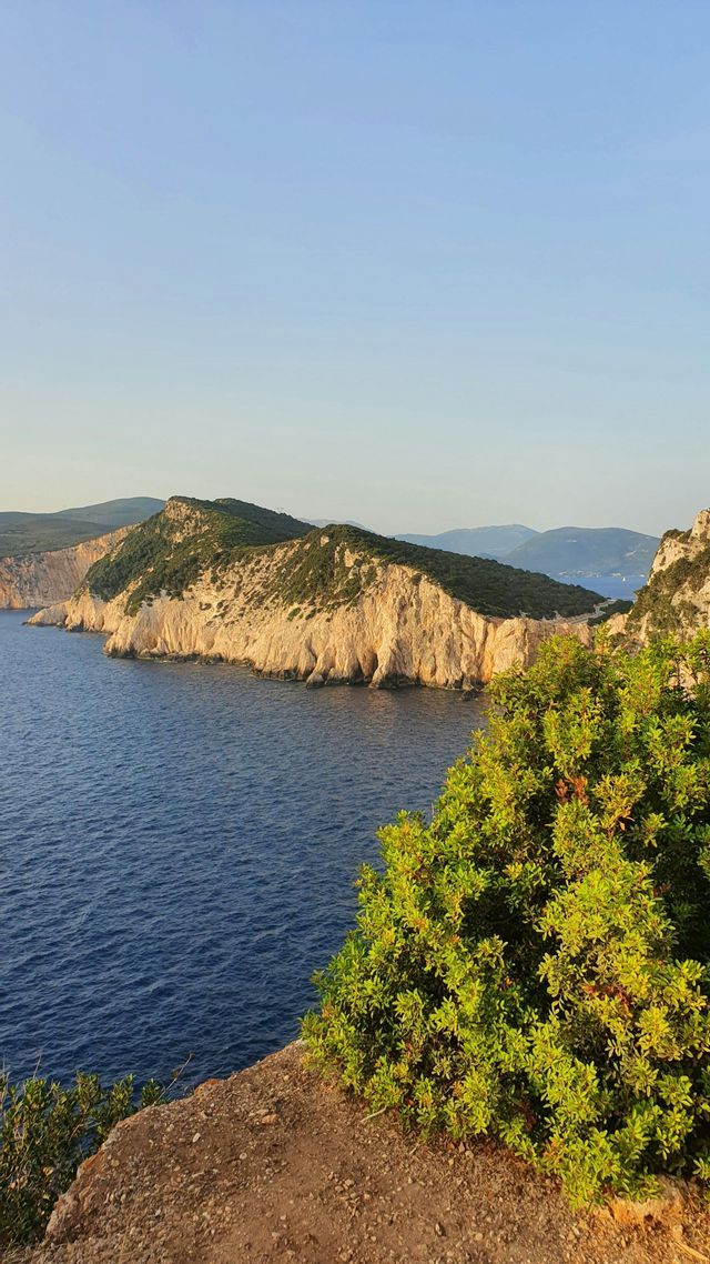 Una vista da una scogliera mostra una penisola rocciosa coperta di alberi verdi che si estende in un mare calmo e blu sotto un cielo sereno.
