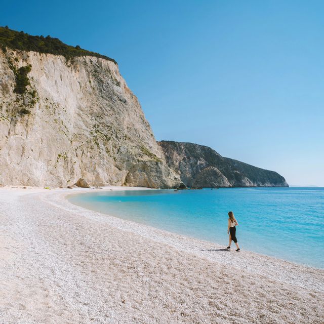 Una donna cammina su una spiaggia di ciottoli bianchi accanto all'acqua turchese, alla base di una grande scogliera bianca sotto un cielo azzurro chiaro.