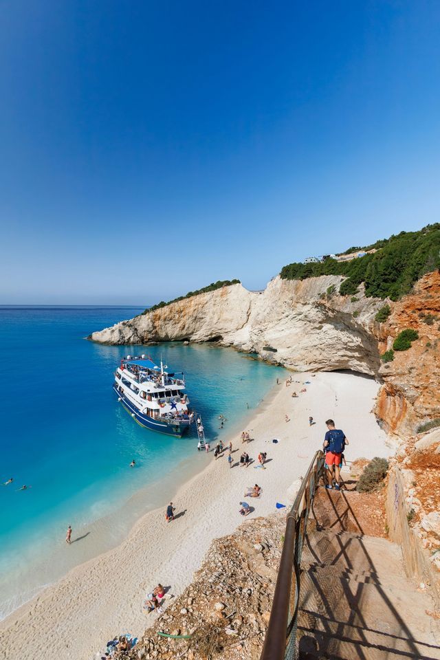 Veduta dall'alto di un gruppo WeRoad che sbarca da una barca su una spiaggia di sabbia bianca, in una cala con acqua turchese e scogliere.
