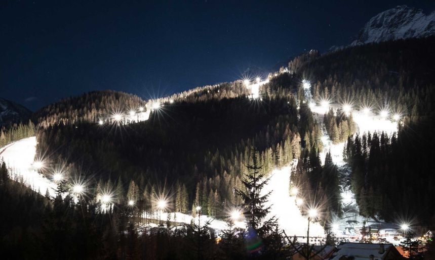 Luci brillanti illuminano una pista da sci innevata che si snoda attraverso una fitta foresta su una montagna di notte sotto un cielo stellato.
