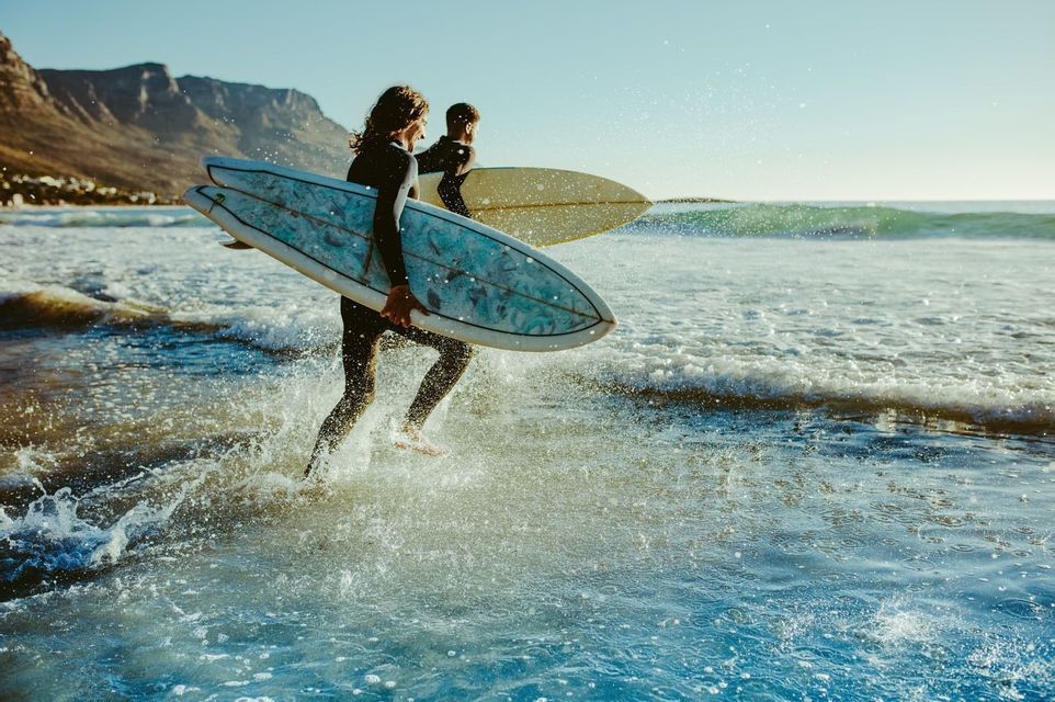 Due surfisti in muta, con le tavole al braccio, corrono nell'acqua bassa dell'oceano, creando schizzi sotto il sole.