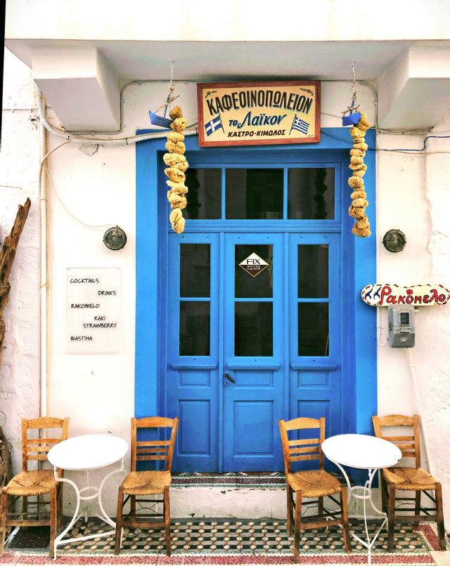 L'ingresso di un caffè in stile greco con una porta blu brillante, spugne marine appese e tavolini con sedie in legno all'esterno.