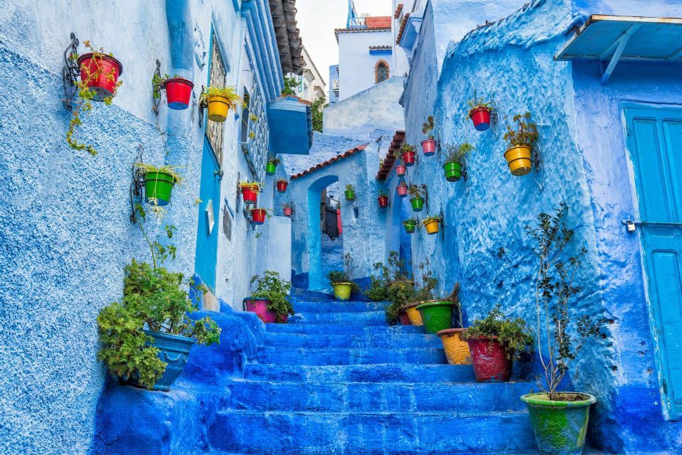 Un callejón estrecho con escaleras, donde las paredes y los escalones están pintados de azul brillante y decorados con macetas de flores coloridas.