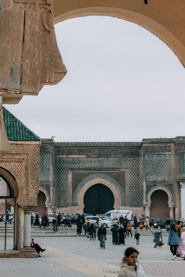 Vista attraverso un arco di pietra, una folla di persone attraversa una piazza cittadina davanti a un grande portale storico con elaborate maioliche.
