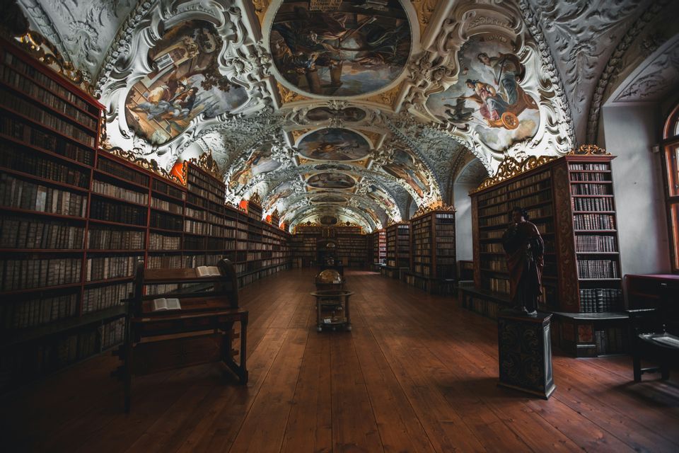 El largo pasillo de una biblioteca histórica con estanterías de madera del suelo al techo y un techo abovedado ornamentado con frescos.