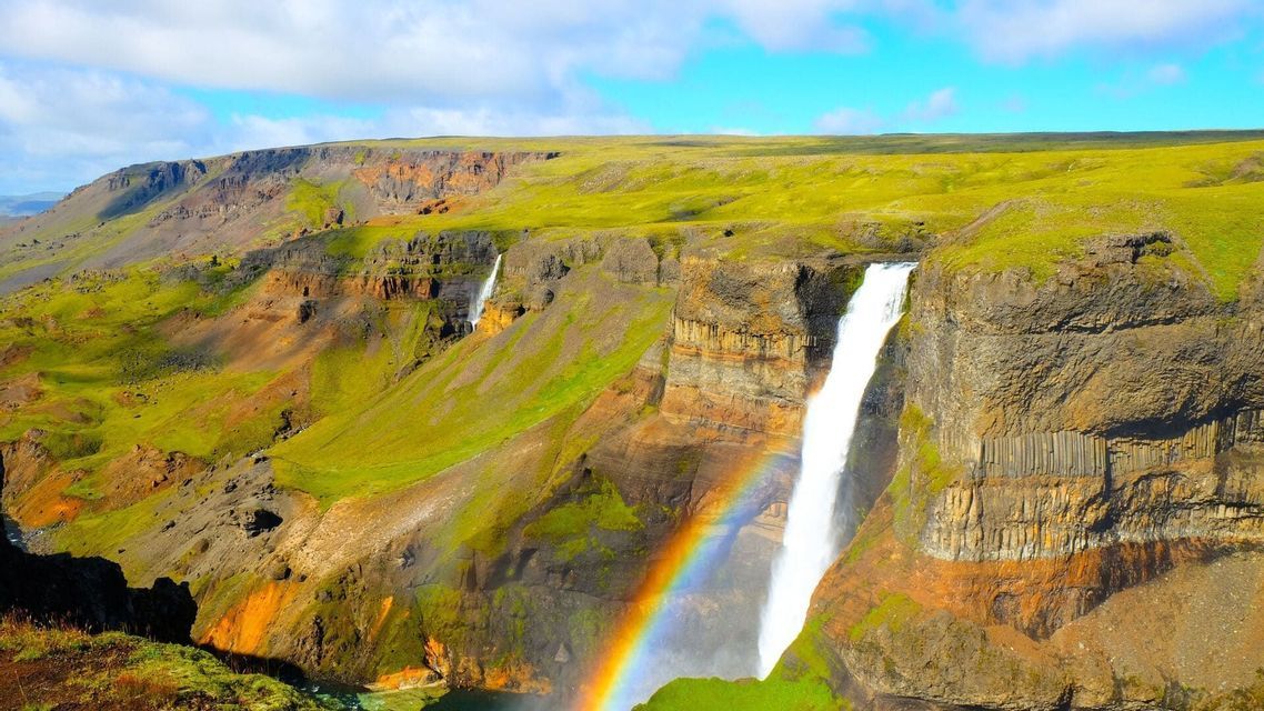 Ein lebendiger Regenbogen spannt sich durch den Nebel eines mächtigen Wasserfalls, der in eine weite, grüne Schlucht mit felsigen Klippen stürzt.