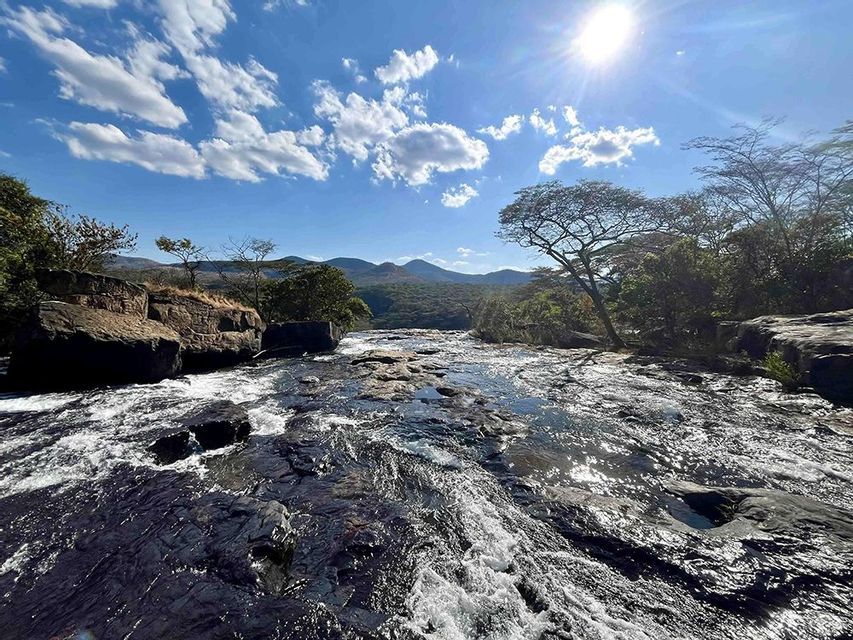Un fiume ampio e roccioso scorre rapidamente sotto un sole splendente e un cielo azzurro nuvoloso, con colline boscose in lontananza.