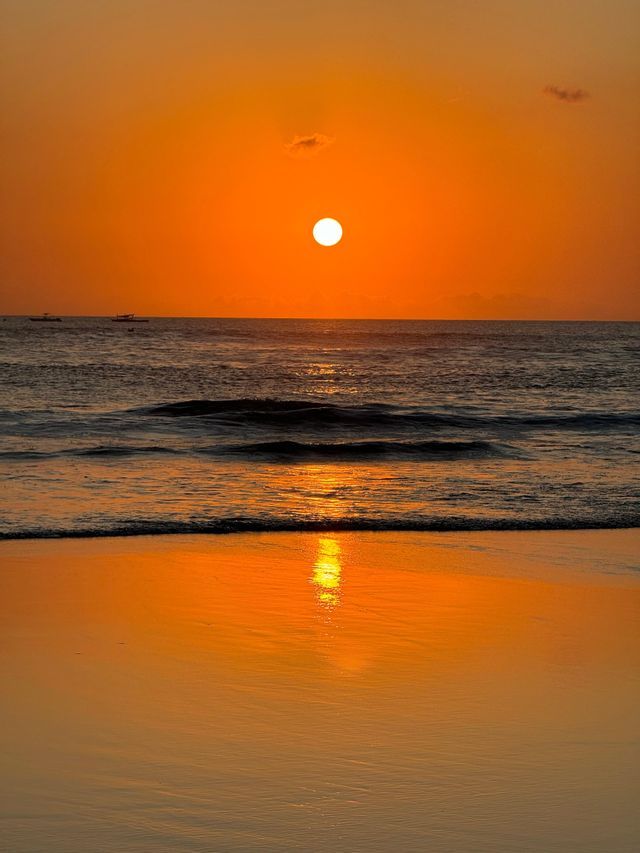 Il sole al tramonto brilla in un cielo arancione, la sua luce si riflette sull'oceano e sulla sabbia bagnata di una spiaggia.