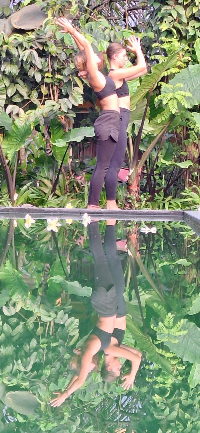 Due donne in posa yoga accanto a una piscina riflettente circondata da una lussureggiante vegetazione verde, creando un riflesso simmetrico nell'acqua.