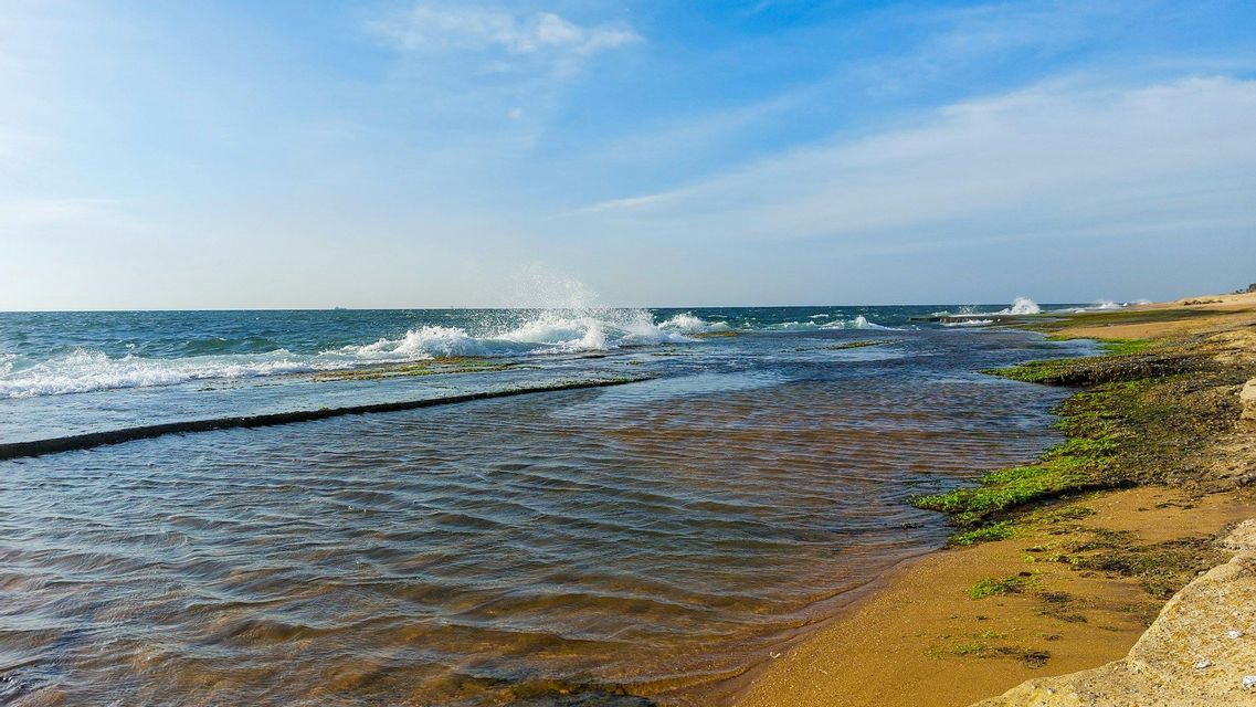 Onde dell'oceano si infrangono su una costa rocciosa e coperta di muschio, accanto a una spiaggia sabbiosa, sotto un cielo parzialmente nuvoloso.