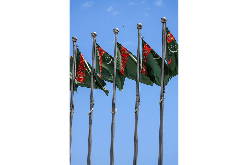 Una fila di bandiere del Turkmenistan che sventolano su aste d'argento contro un cielo azzurro chiaro.