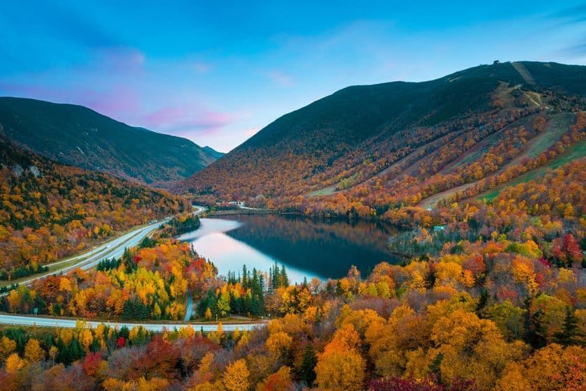 Una vista aerea di una strada tortuosa accanto a un lago circondato da montagne con alberi autunnali colorati.
