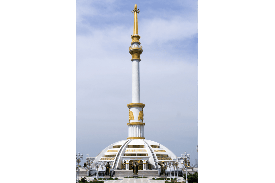 Un alto monumento bianco, con guglia e dettagli dorati, si erge su una base a cupola in un'ampia piazza sotto un cielo nuvoloso.