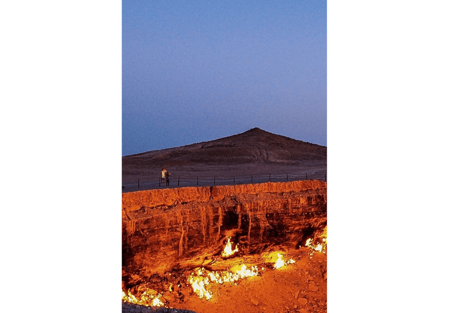 Due persone di un viaggio di gruppo WeRoad si trovano accanto a una ringhiera, guardando un grande cratere di gas infuocato nel deserto al tramonto.