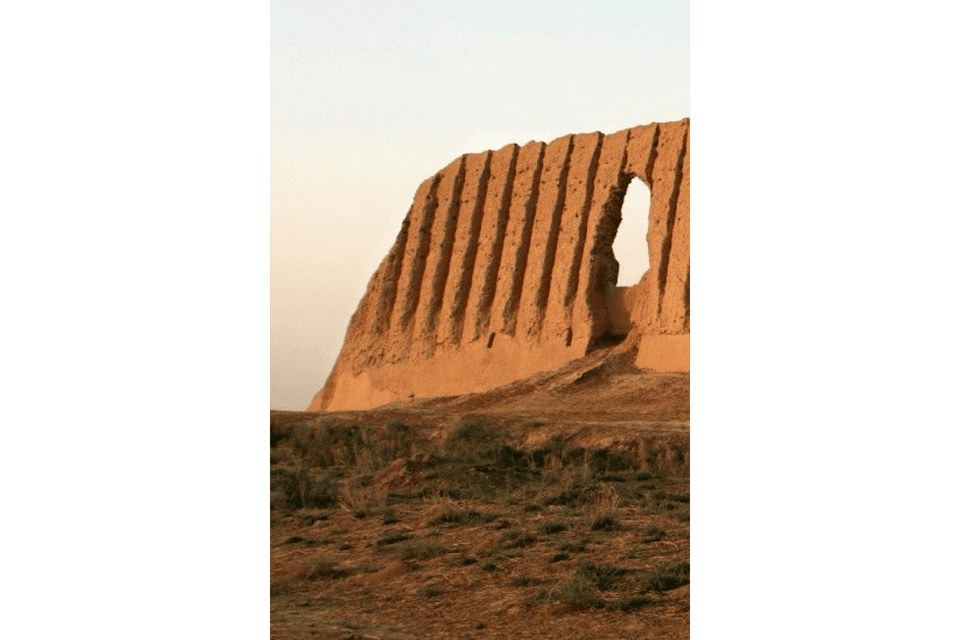 Una rovina di muro in mattoni di fango ondulato con una grande apertura si erge su una collina secca ed erbosa contro un cielo pallido e velato.