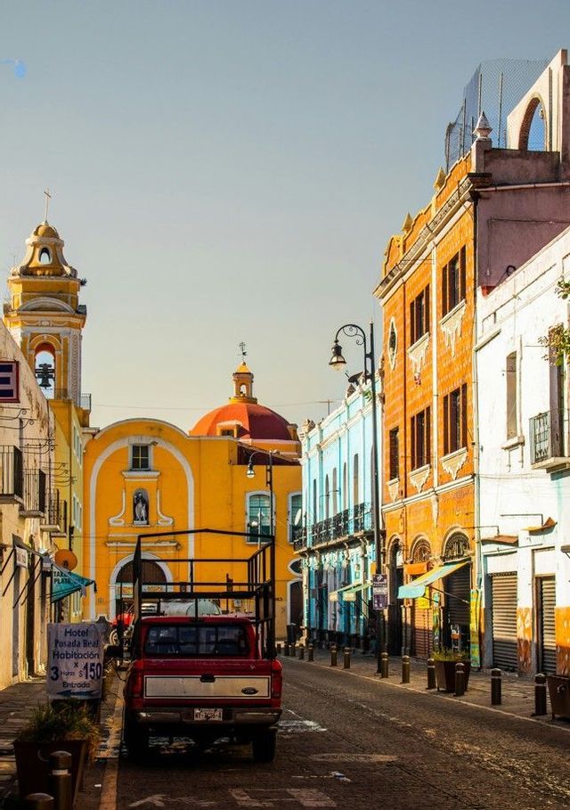 Una camioneta roja aparcada en una calle estrecha flanqueada por edificios coloridos, que conduce hacia una iglesia amarilla con cúpula roja.