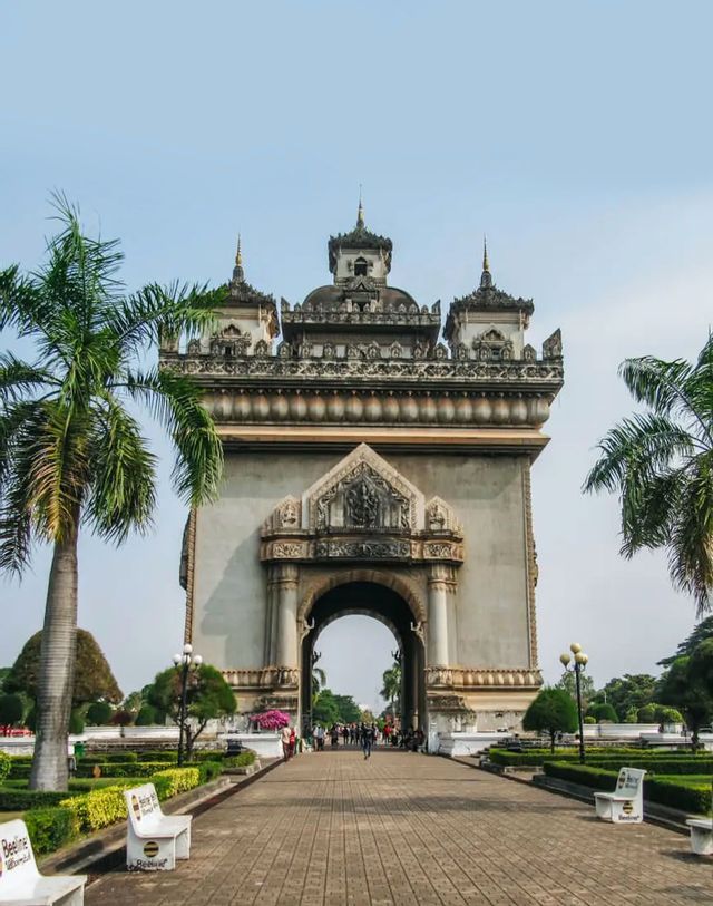 Un arco trionfale in pietra con intagli intricati si erge su un sentiero lastricato, circondato da giardini con palme e panchine bianche.
