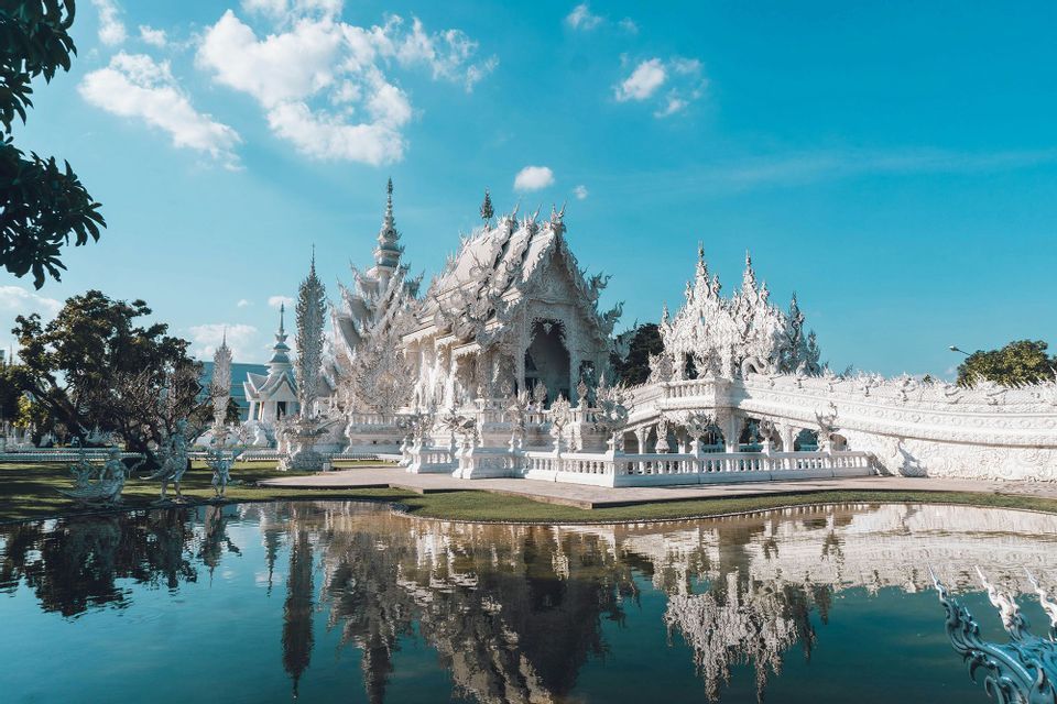 Un tempio bianco ornato con guglie intricate che si riflette in uno specchio d'acqua calmo sotto un cielo azzurro con nuvole.