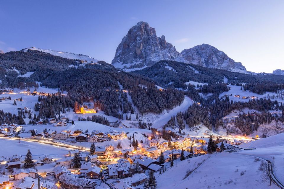 Un piccolo villaggio con luci scintillanti è incastonato in una valle innevata, ai piedi di un'imponente montagna ricoperta di neve, al crepuscolo.