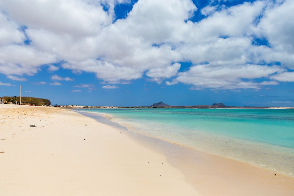 Une large plage de sable blanc rencontre une eau turquoise limpide sous un ciel bleu parsemé de nuages blancs.