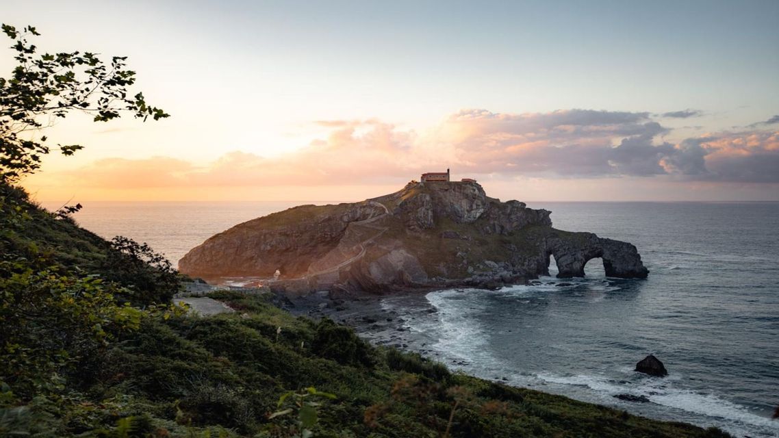 Un isolotto roccioso con un edificio storico in cima, visto da una scogliera verde mentre il sole tramonta sull'oceano.
