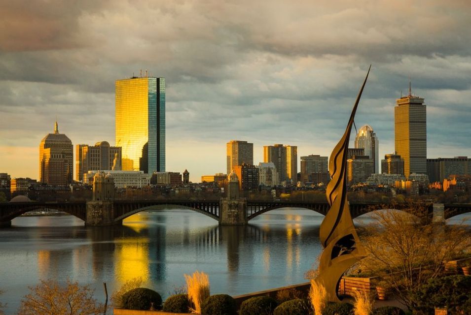 Uno skyline cittadino con grattacieli che riflettono la luce dorata del tramonto su un fiume con un ponte.