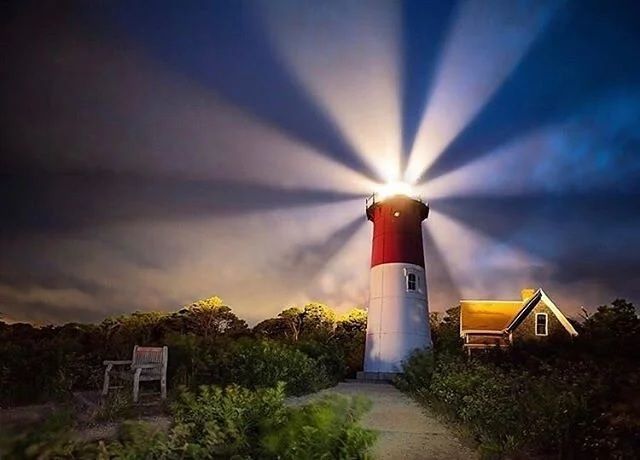 Un faro rosso e bianco proietta potenti fasci di luce attraverso un cielo notturno scuro e nuvoloso, con un piccolo cottage e alberi nelle vicinanze.