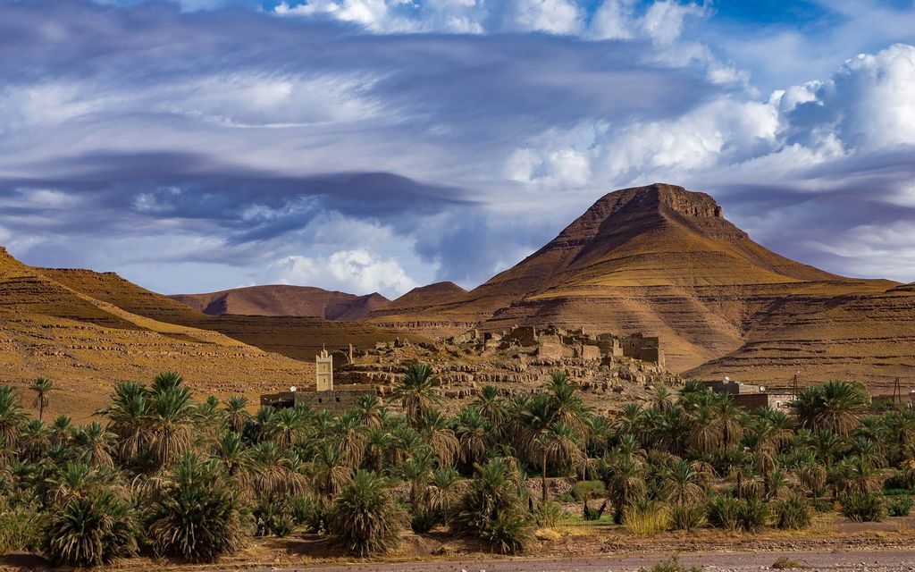 Un antico villaggio e minareto incastonati in una lussureggiante oasi di palme, ai piedi di aride montagne stratificate sotto un cielo nuvoloso.