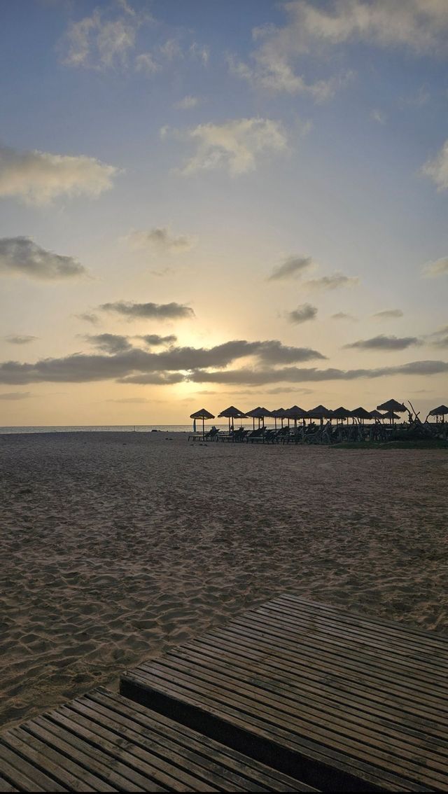 Una passerella di legno su una spiaggia sabbiosa, con una fila di ombrelloni e lettini sagomati contro il tramonto sull'oceano.