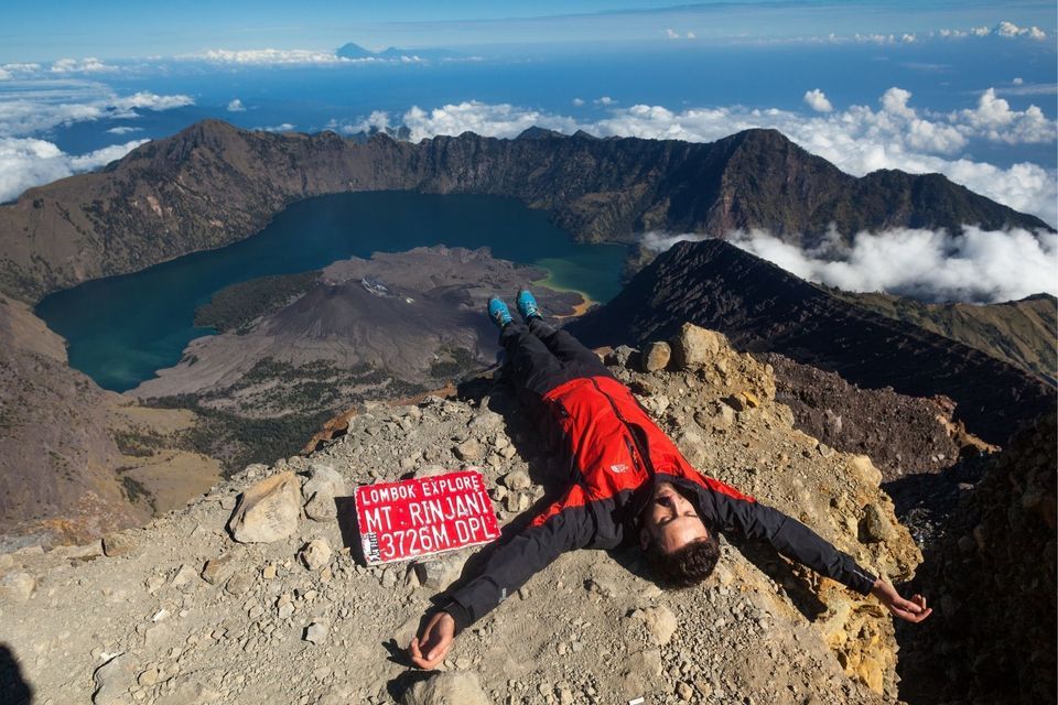 Ein Mann in einer roten Jacke ruht auf einem felsigen Berggipfel und blickt auf einen riesigen Vulkankrater mit einem See darin.