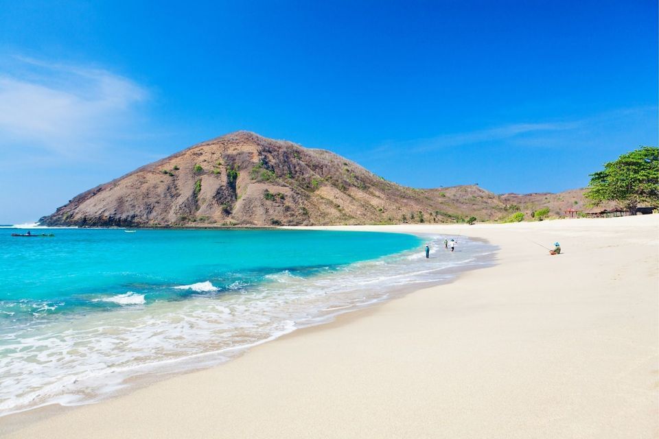Ein weißer Sandstrand grenzt an eine Bucht mit türkisfarbenem Wasser, im Hintergrund ein großer, karger Hügel unter strahlend blauem Himmel.