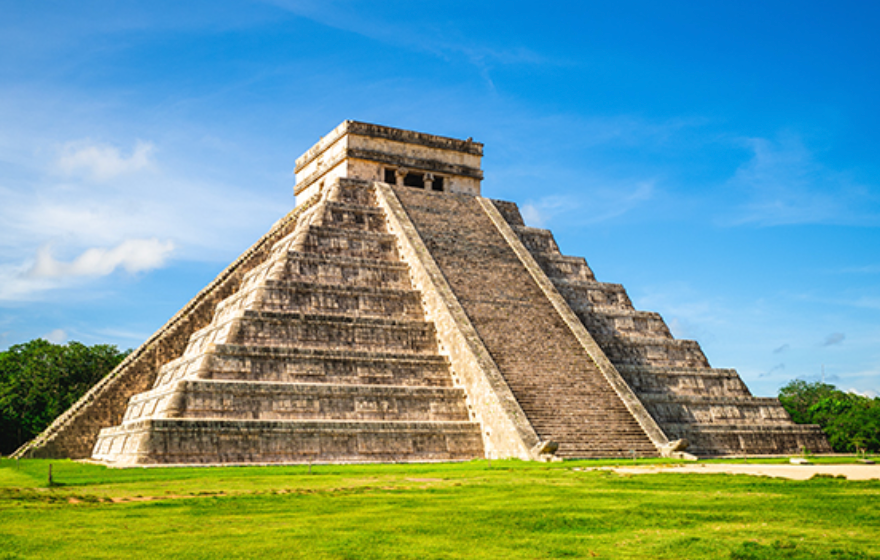 A large stone step pyramid with steep staircases stands on a grassy field under a bright blue sky.