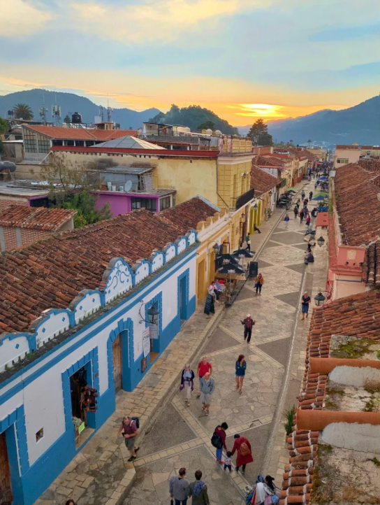 A high-angle view of a colorful town street where people walk among buildings with terracotta roofs as the sun sets behind mountains.