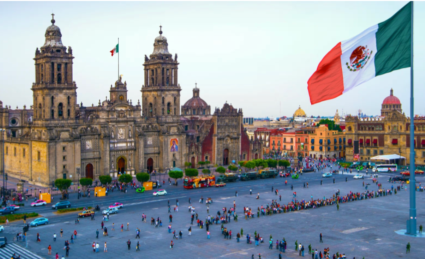 The Mexican flag waves over a bustling city square with a large, historic cathedral in the background.